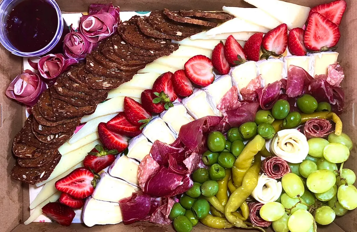 Beautiful grazing board featuring charcuterie-style elements, including crackers, sliced cheese, fresh strawberries, cured meats, green olives, pickled peppers, and grapes, perfect for catering events.