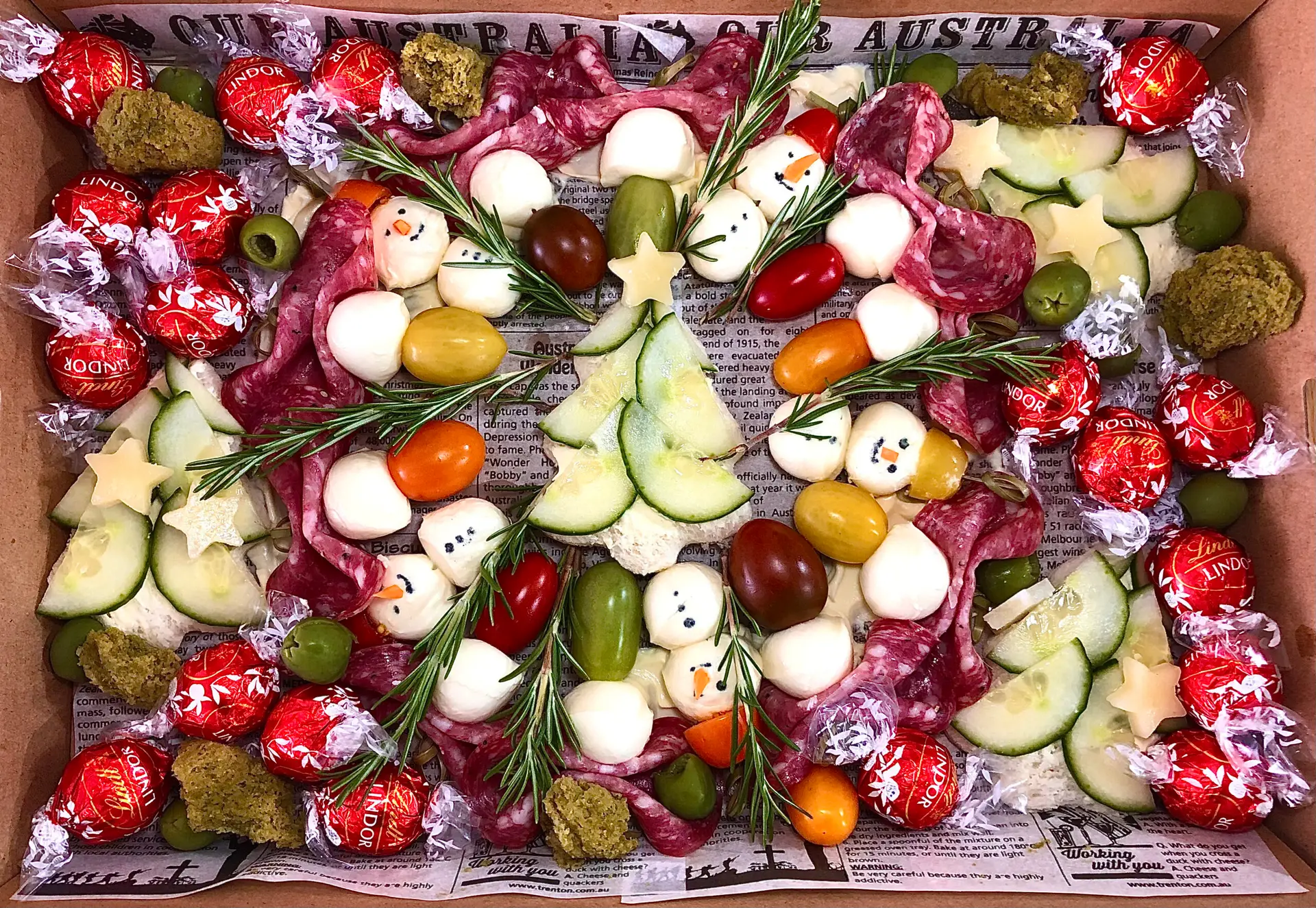 Festive Christmas grazing board with charcuterie-style treats, including snowman mozzarella, cherry tomatoes, cucumber slices, star-shaped cheese, rosemary sprigs, Lindor chocolates, salami, and olives, designed for holiday catering.