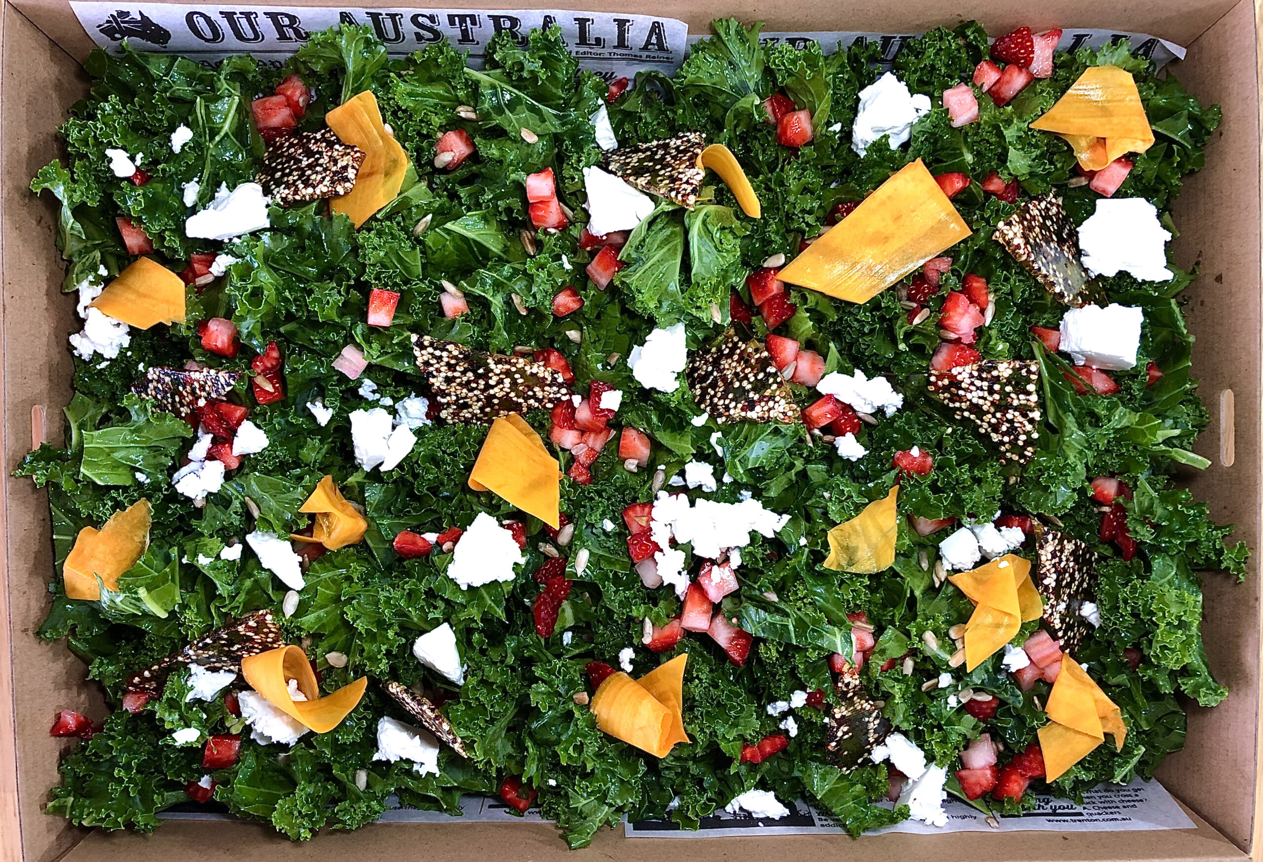 Fresh kale and strawberry salad in a catering box, topped with feta cheese, walnuts, and a light dressing, perfect for gourmet events and healthy meal options.