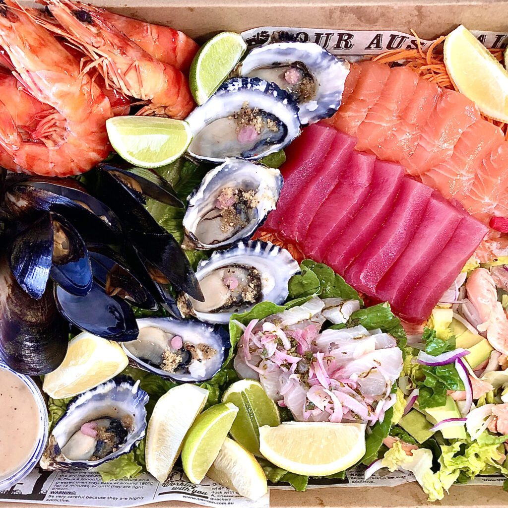 Fresh seafood platter featuring prawns, mussels, oysters, sashimi slices of tuna and salmon, and vibrant salads with lime wedges and salad sauce.