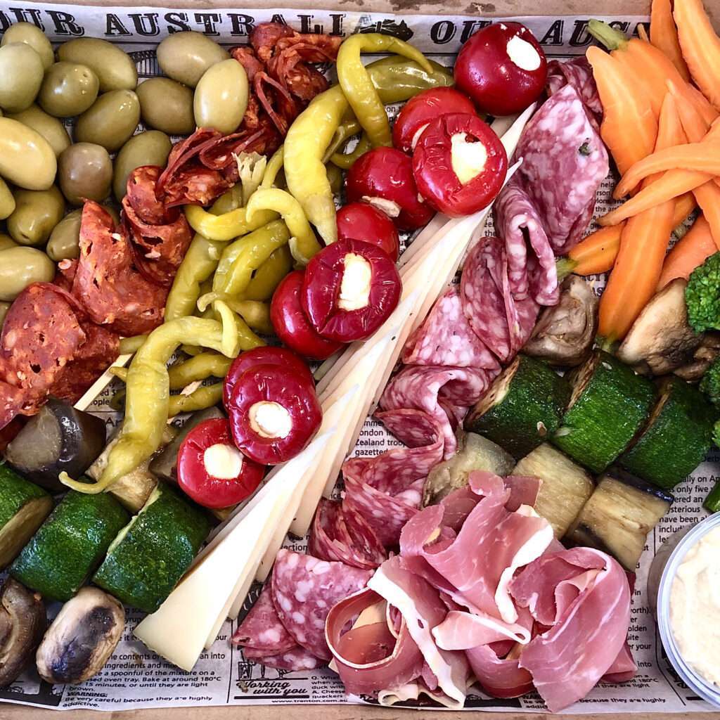 Colorful antipasto platter featuring olives, salami, prosciutto, stuffed peppers, pickled vegetables, roasted zucchini, eggplant, carrots, broccoli, and creamy hummus.