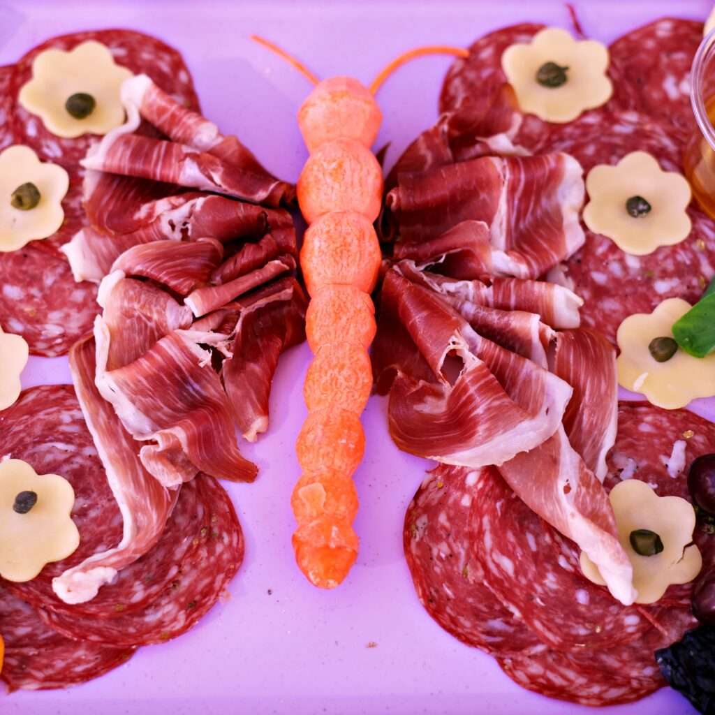 Butterfly-shaped kids charcuterie platter made with prosciutto, salami, cheese flowers, grapes, apricots, and carrot body.