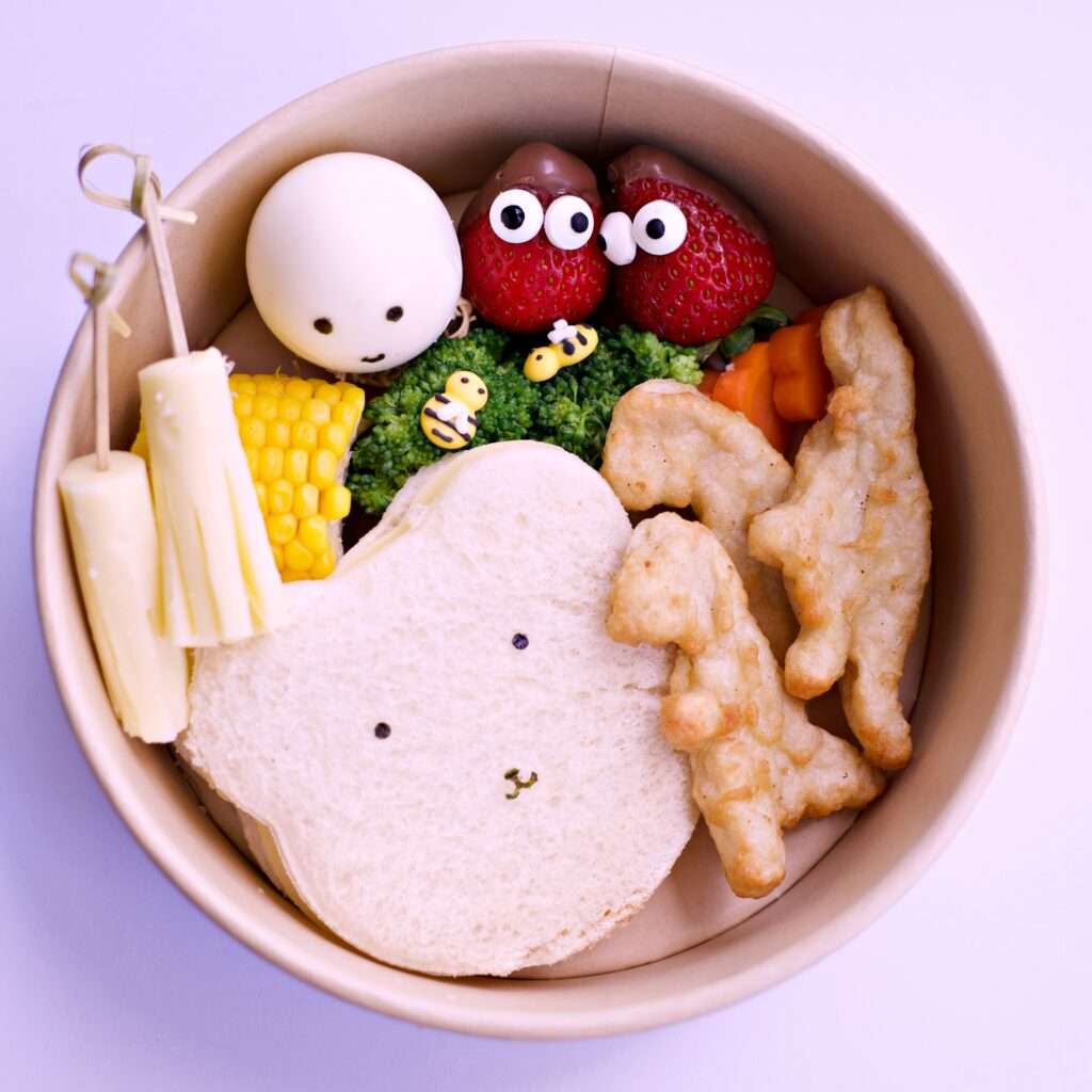 Cute kids lunch box with baby bear sandwich, dinosaur nuggets, cheese stick, corn, boiled egg, broccoli, and strawberry decoration.